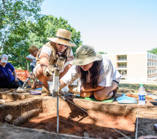 Two Clemson students in wide-brimmed hats measure the depth of a dig they're conducting on campus in front of the Shoeboxes.