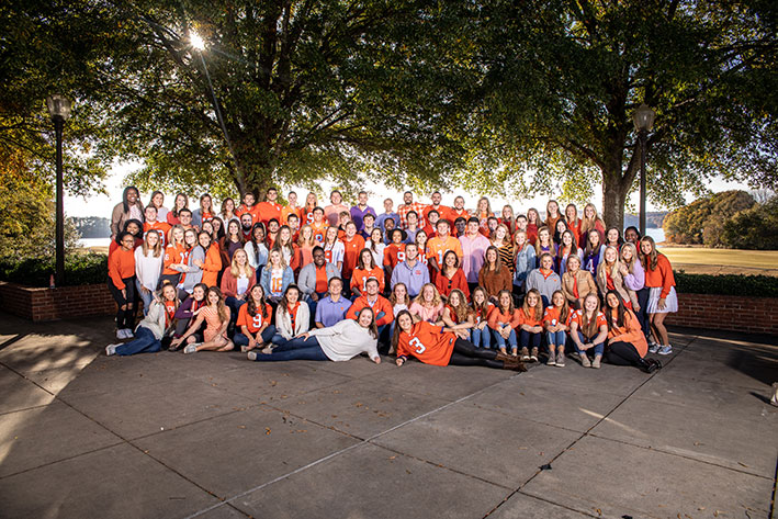 A large group of student tour guides pose together, wearing varying shades of orange in a shaded spot beside Lake Hartwell.