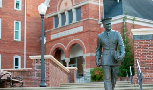 A statue shows a Clemson cadet holding a book striding from campus toward Bowman Field.
