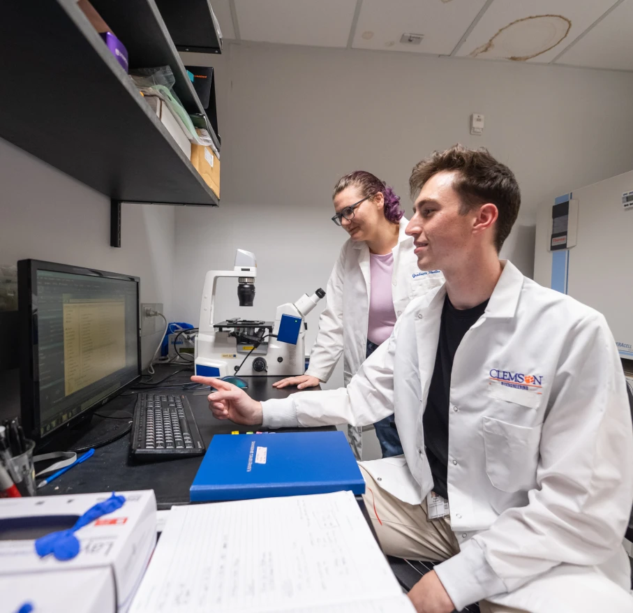 A male student sits in front of a table with a computer, notebooks and microscope, and a female professor stands next to him in a laboratory setting.