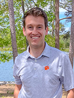 A smiling man standing outdoors near a lake, wearing a gray polo shirt with an orange Clemson logo.