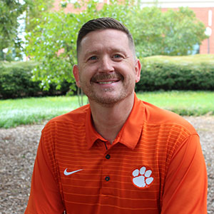 Smiling man in a Clemson University orange polo shirt sitting outdoors with greenery in the background.
