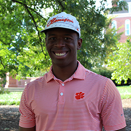 Smiling man wearing a white cap and a red-and-white striped polo shirt outdoors near a campus building.