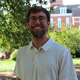 Portrait of a smiling man wearing glasses and a light polo shirt outdoors with trees and a building in the background.