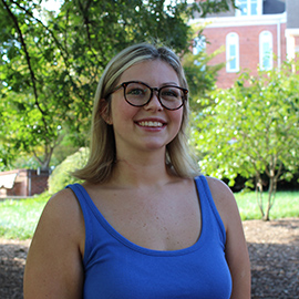 Smiling person wearing glasses and a blue tank top outdoors near trees and a campus building.