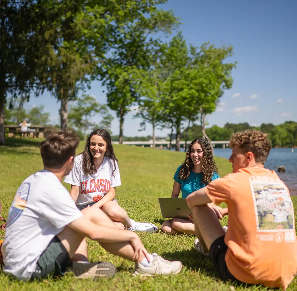 A group of students sit and talk on a grass lawn in front of lake Hartwell with Clemson's main campus in the background