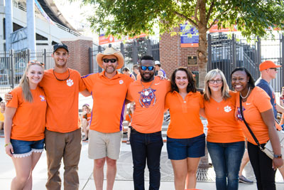 Seven Clemson fans wearing orange shirts pose together outside Memorial Stadium.