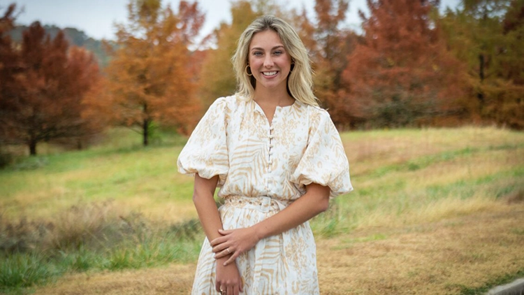 A female student wearing a white dress with a beige pattern of stripes and spots stands in a grassy field with red-leafed trees in the background