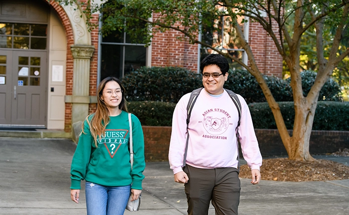 A male student wearing a pink sweatshirt and dark gray pants walks with a female student wearing a teal sweatshirt and jeans outside a brick academic building.