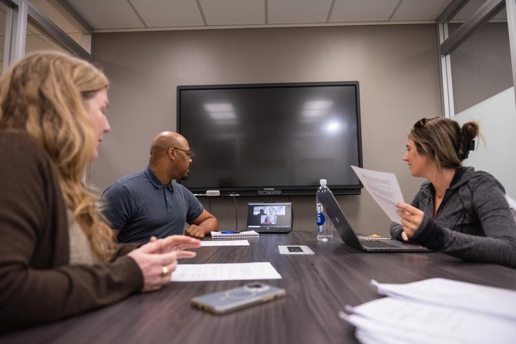 A man and two women sit at a table with paper and laptops in front of them and look at a laptop screen at the end of the table that is running a video conference session.