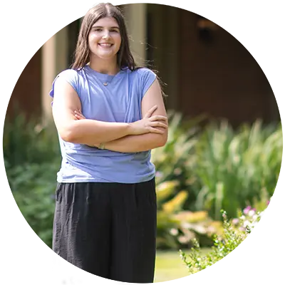 : A female student wearing a light blue sleeveless top poses with her arms crossed in front of an renovated historic home surronded by vegitation