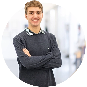 A male student wearing a gray-blue sweater and khaki pants poses inside an architecture studio space. 