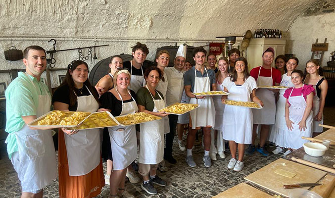 A line of study abroad students hold up pans of various types of pasta in a large ornate kitchen.