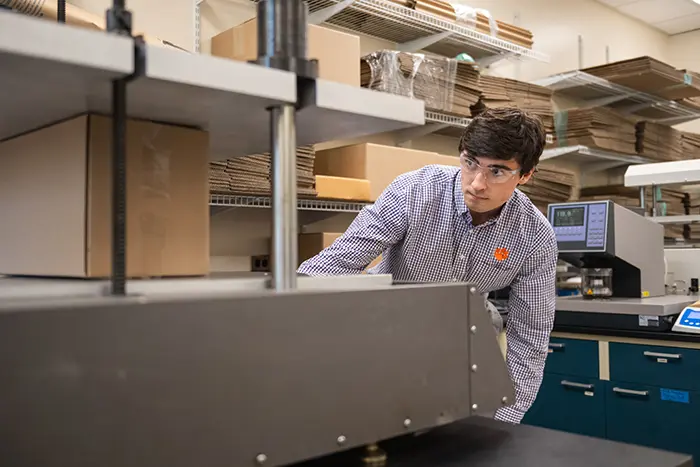 A male student wearing a purple gingham dress-shirt and safety glasses measures the structural strength of a cardboard box on a hydraulic press.