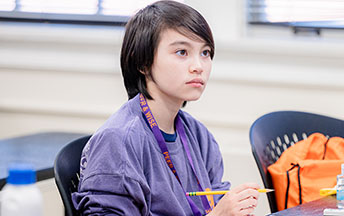 A young student holding a pencil looks up from their desk towards the front of the room.