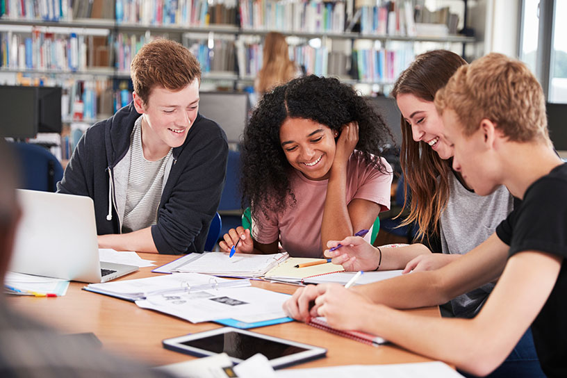 Students smile while studying at a table scattered with notebooks and paper.