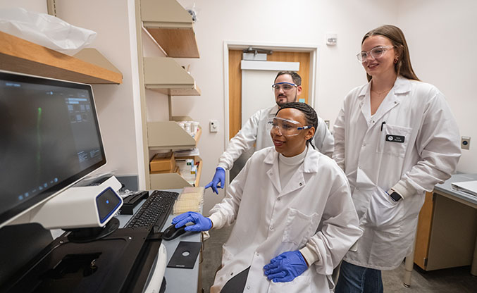 A male and female student overlook their female professors shoulder to view a monitor. All are wearing white lab coats, exam gloves and protective eyewear.