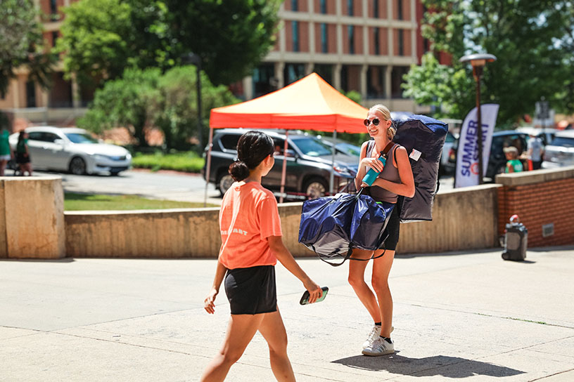 Two female students cross paths as one carries a large duffle bag in front of a Summer Start banner.