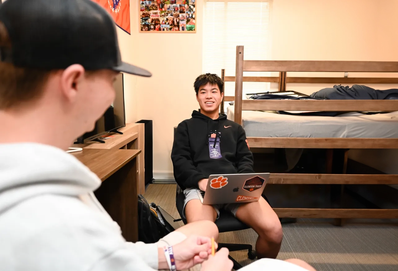 A male sudent and a friend hang out in a residence hall, sitting with their laptops out beside a bed, TV and desk. 