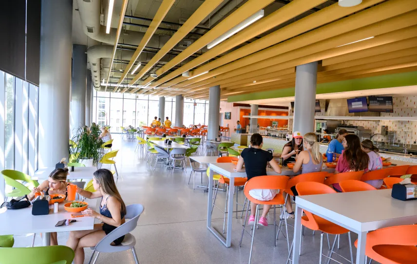 Students sit and eat in a dining hall with window-to-celiing walls, slate grey flooring, orange chairs and wooden accents. 