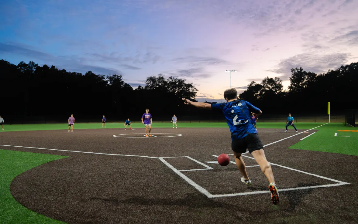 Students play kickball on a pristine baseball pitch at sunset.