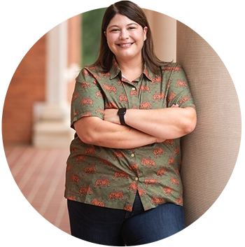 A woman wearing blue jeans and an olive short-sleeved button-down shirt with a pattern of orange tigers stands with her arms folded next to a pillar of a building. 