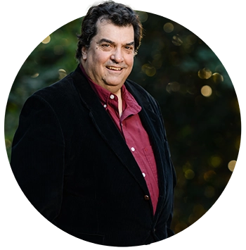 : A man wearing a burgundy button-down Oxford shirt and a black blazer poses in front of a blurred background of trees on campus. 