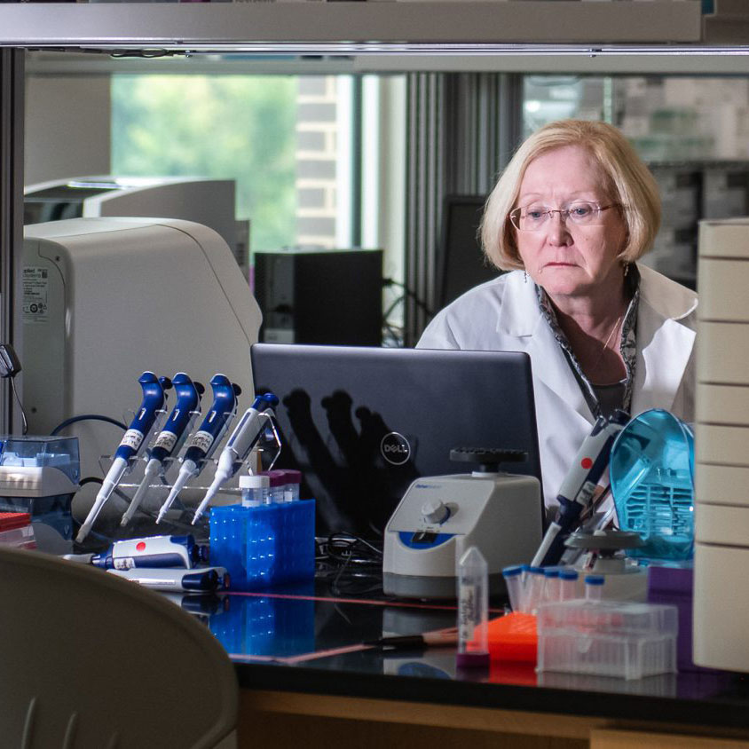 Dr.Trudy Mackay sits in front of a computer in lab surrounded by research equipment