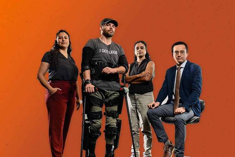 Two female professors standing and a male professor sitting on a stool next to a man whose legs and arms are outfitted with an exoskeleton device.