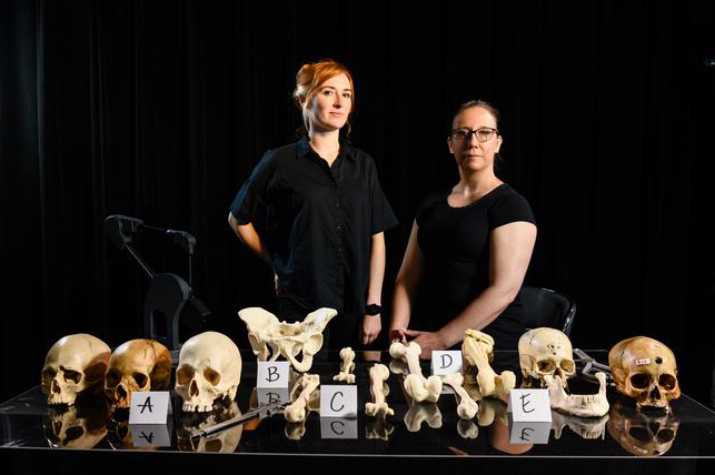 Two women wearing black tops pose behind a collection of instructional models of skeletons, including skulls and bones, displayed on a table.