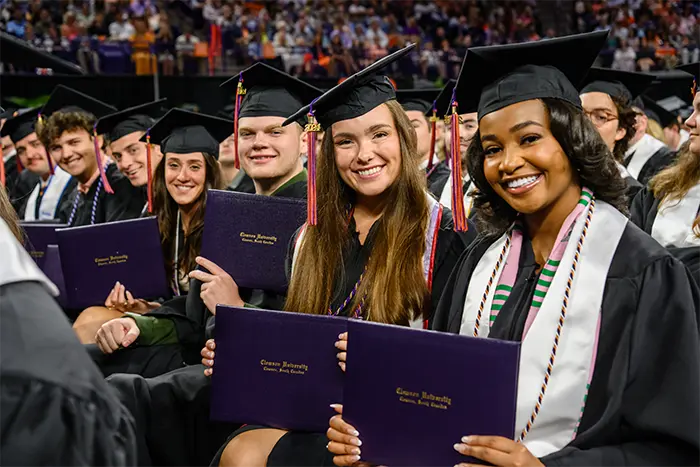 Seven seated students sitting in a row during Commencement wear black graduation caps and
gowns and smile while showing the outside covers of their Clemson University diplomas.