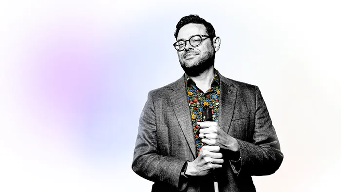 A photo illustration of a man wearing a floral shirt and a blazer and holding a white cane — all edited to be in black and white except for the multicolored floral shirt — with an added pink-and-purple blurred light flare effect to his left.