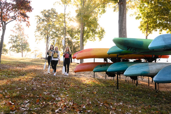 three students in lifejackets walk alongside canoes and kayaks