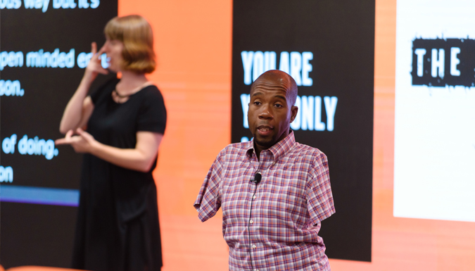 African American man without both arms, speaking to an audience with a blurry digital display and sign language interpreter in the background