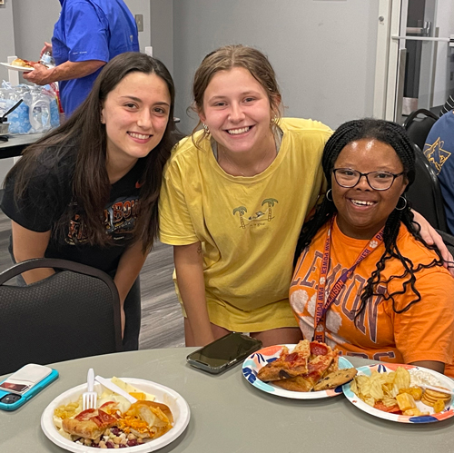 3 female students looking at the camera and embracing around a dinner table for friendsgiving