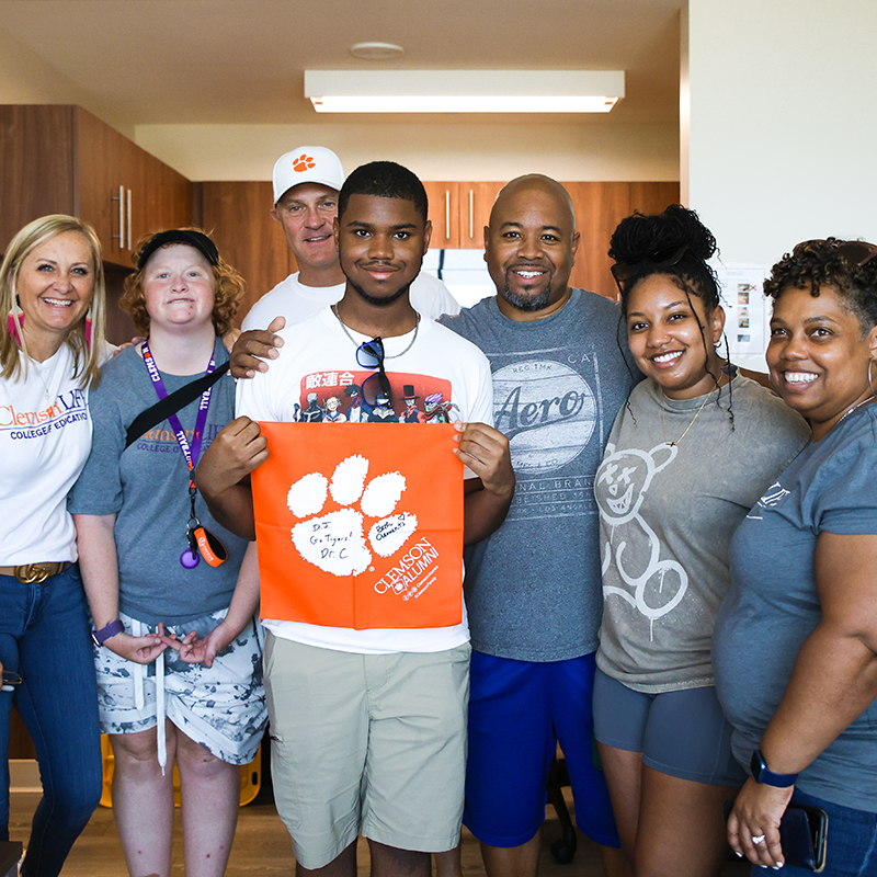 a students holding an orange clemson tiger rag poses in a student housing apartment with his family and president clements family.