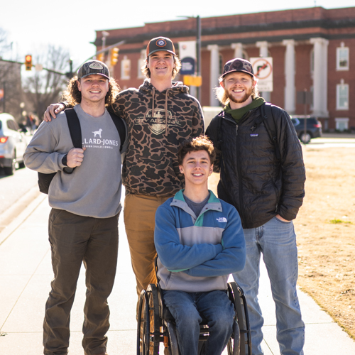 group of male students smiling at the camera 3 standing and one in a wheelchair