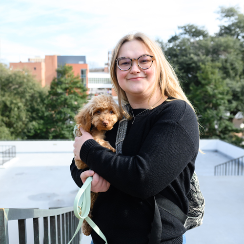 female student smiling, facing the camera and holding a small, cream colored dog