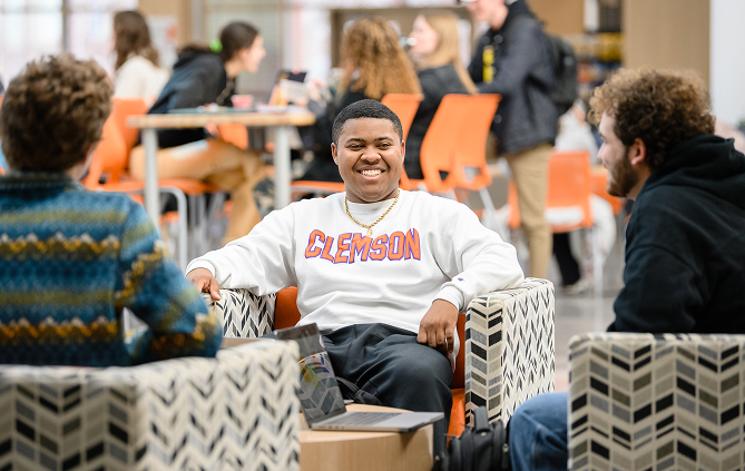 A male student sits in a lounge chair across from two other male students.