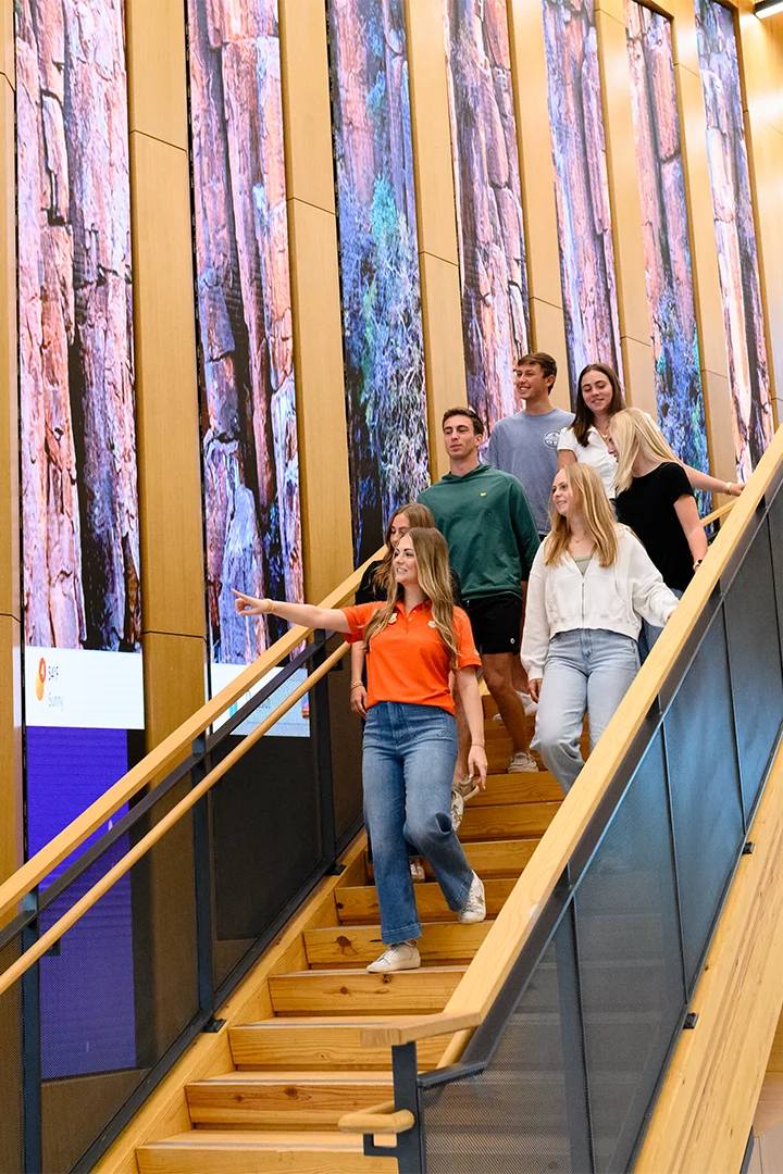 A group of students ascending stairs inside a building, engaged in conversation and carrying backpacks.