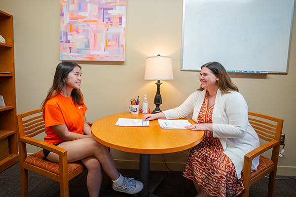 Two women engage in a conversation at a round table in a cozy office setting, with one pointing to a document while the other listens attentively.