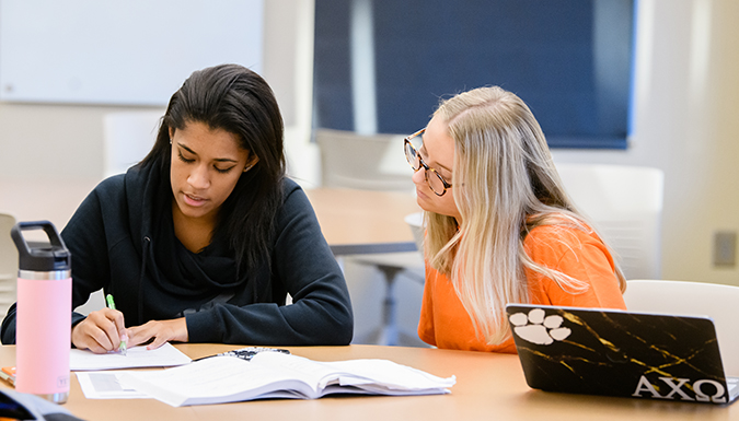 Two students engaging in conversation at a table in a classroom with a laptop and a relaxed atmosphere.