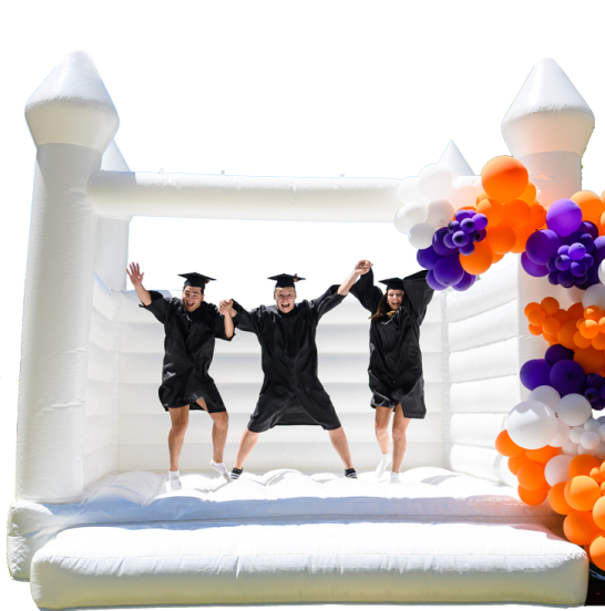 Three graduates in cap and gowns jump in a white bounce house decorated with orange and purple balloons. 