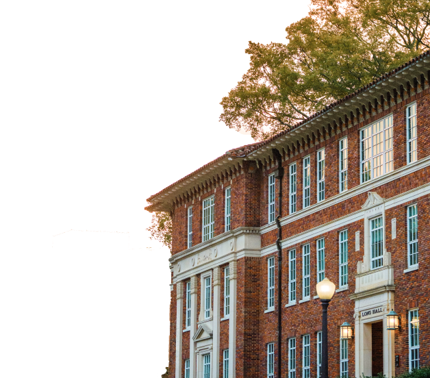 A tree peaks over the top of a three story red brick building with many windows and a series of small columns surrounding the main entrance.