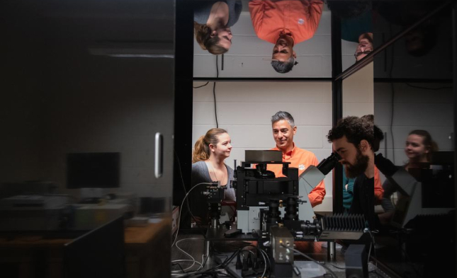 A researcher looks into a microscope lens in a darkroom environment while other look on.