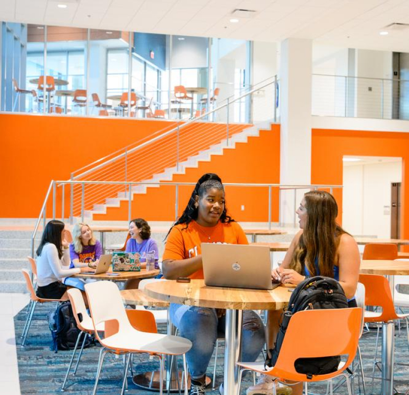 Female students sit around round tables with their laptops while talking with one another