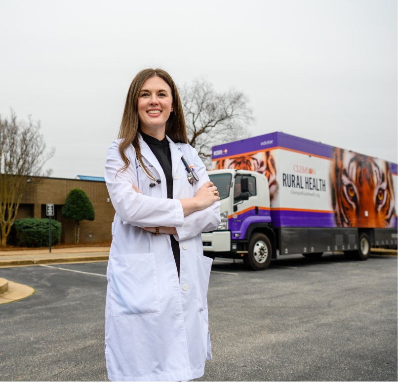 Caitlin Kickham smiles outside of a mobile health clinic, wearing her scrubs and white nursing coat. 