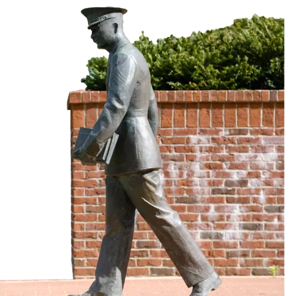 The statue of the senior cadet walking from Military Heritage Plaza onto Bowman Field stands in front of a brick wall and green foliage. 