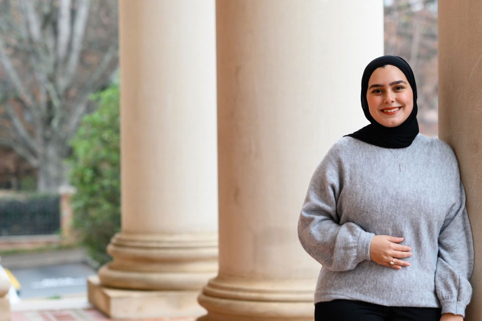 Portrait of a smiling woman wearing a hijab and business casual dress, leaning against a column in front of Sikes Hall. 