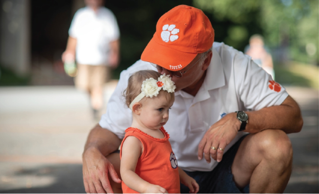 A grandfather crouches to kiss his toddling granddaughter on the head. Both wear Clemson gear.